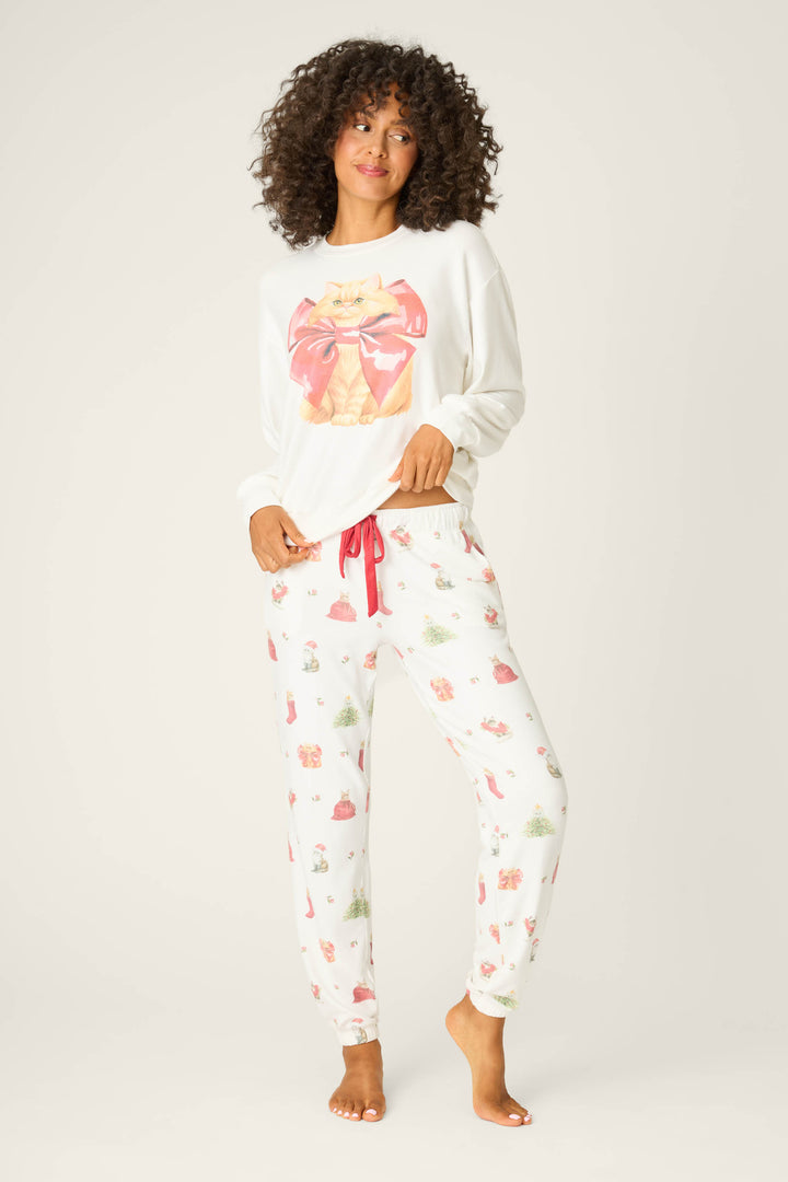 A woman with curly hair stands barefoot against a plain background, wearing a white long-sleeve pajama top and P.J. Salvage Feline Festive Cat Print Banded Pants with a red drawstring and colorful cat patterns.