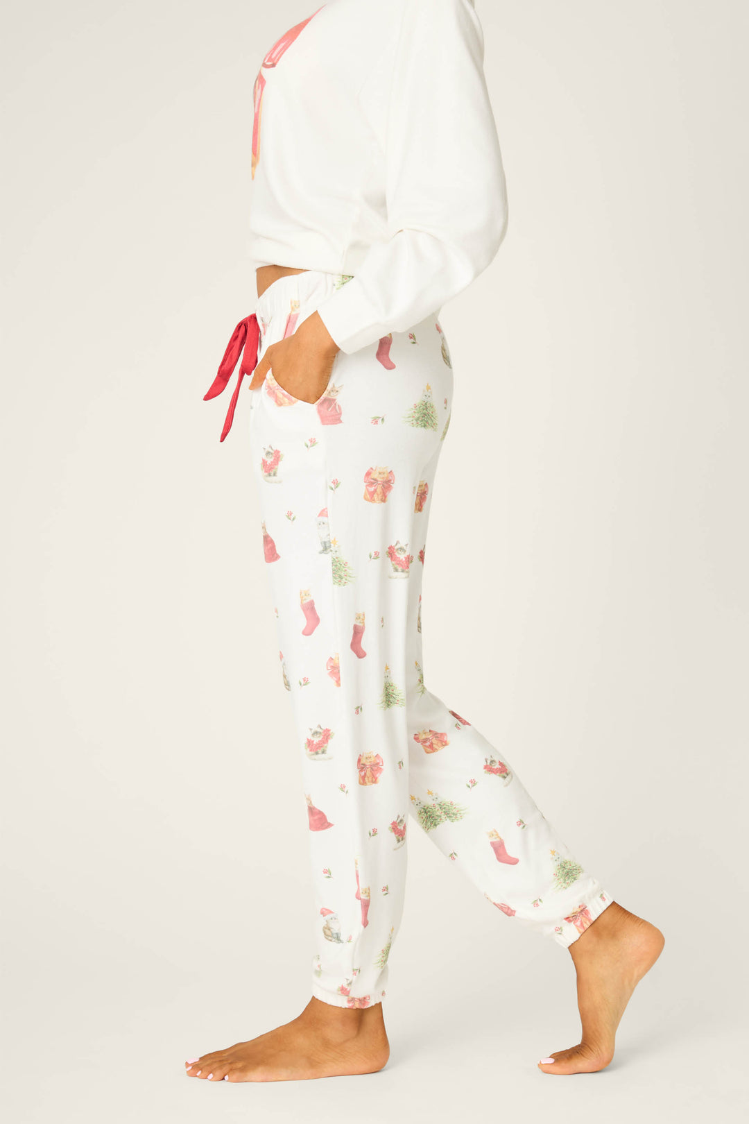 A person is standing sideways wearing P.J. Salvage Feline Festive Cat Print Banded Pants with a red drawstring and side pockets, paired with a matching long-sleeve top. The person is barefoot against a plain background.