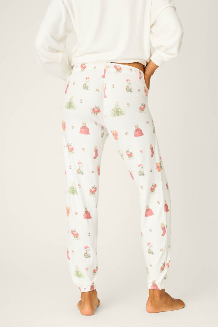A person stands barefoot facing away, wearing P.J. Salvage Feline Festive Cat Print Banded Pants featuring a holiday cat print; the matching white top is partially visible against a plain background.