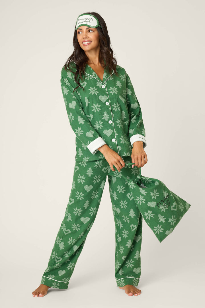 A woman stands barefoot against a white background wearing P.J. Salvage's 4-Piece Tree & Heart Print Flannel PJ Set, featuring green pajamas, a matching drawstring bag, and a coordinating green and white sleep mask.