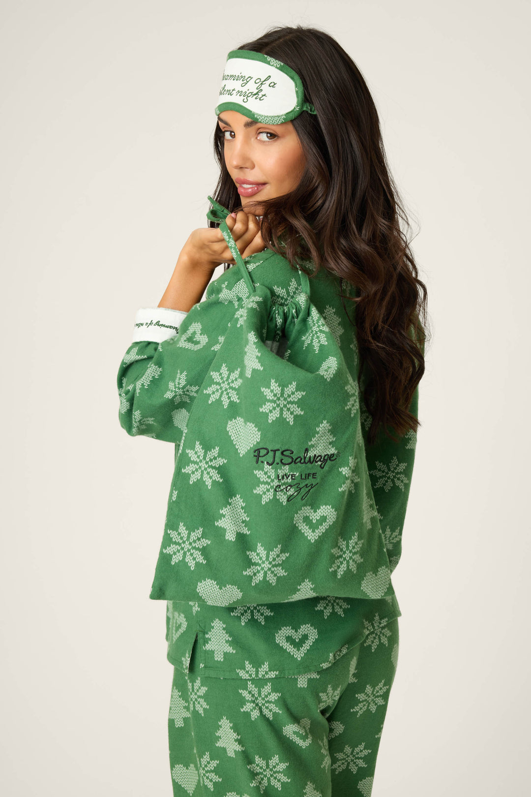 A woman wears the P.J. Salvage 4-Piece Tree & Heart Print Flannel PJ Set and matching sleep mask, holding a branded drawstring bag. She faces backward, looking over her shoulder.