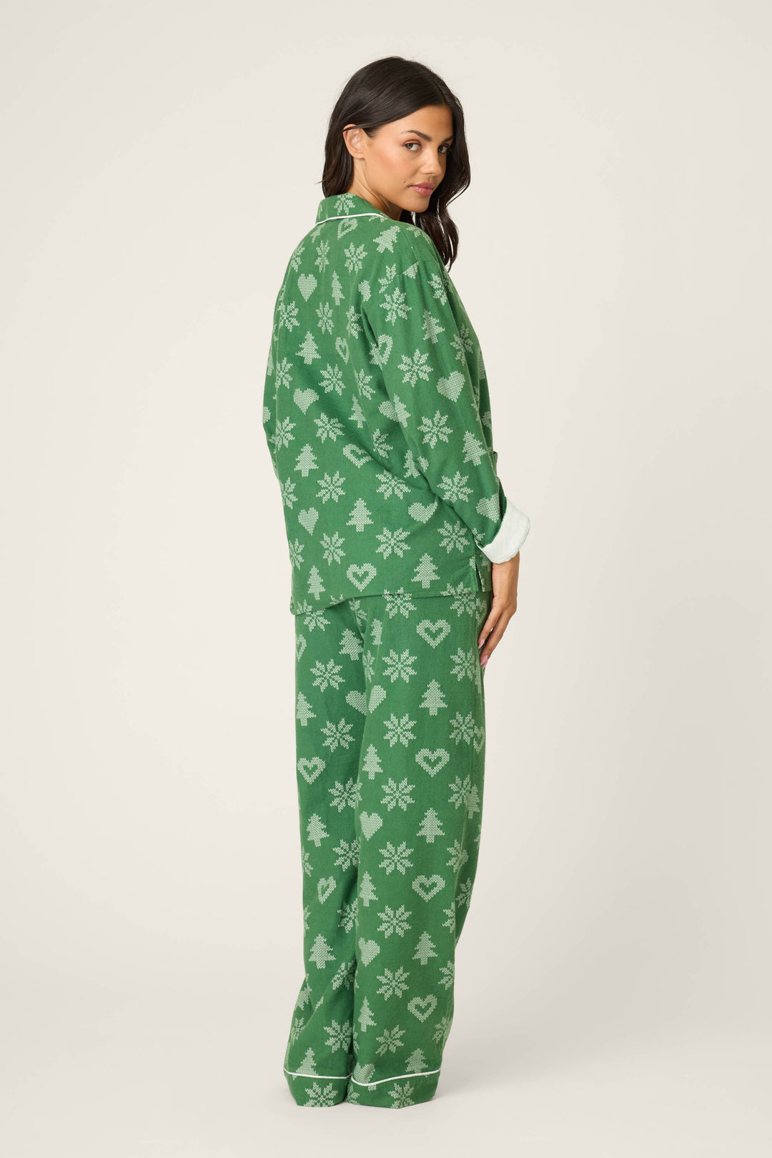 A woman looks over her shoulder wearing the P.J. Salvage 4-Piece Tree & Heart Print Flannel PJ Set, featuring green pajamas with a tree and heart pattern. The background is plain and light-colored.