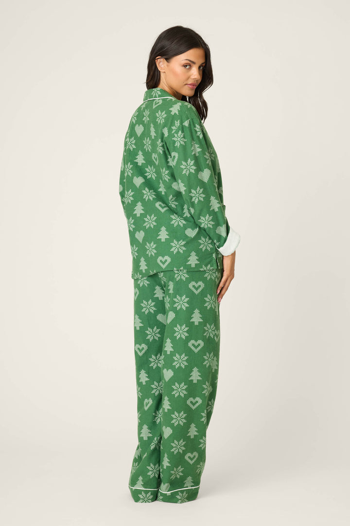 A woman looks over her shoulder wearing the P.J. Salvage 4-Piece Tree & Heart Print Flannel PJ Set, featuring green pajamas with a tree and heart pattern. The background is plain and light-colored.