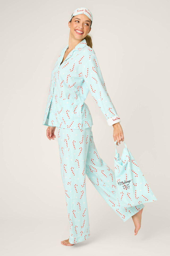 A smiling person stands barefoot against a plain background, wearing the P.J. Salvage 4-Piece Candy Cane Print Flannel PJ Set—light blue pajamas with a red candy cane print, a matching tote bag, and a matching sleep mask.
