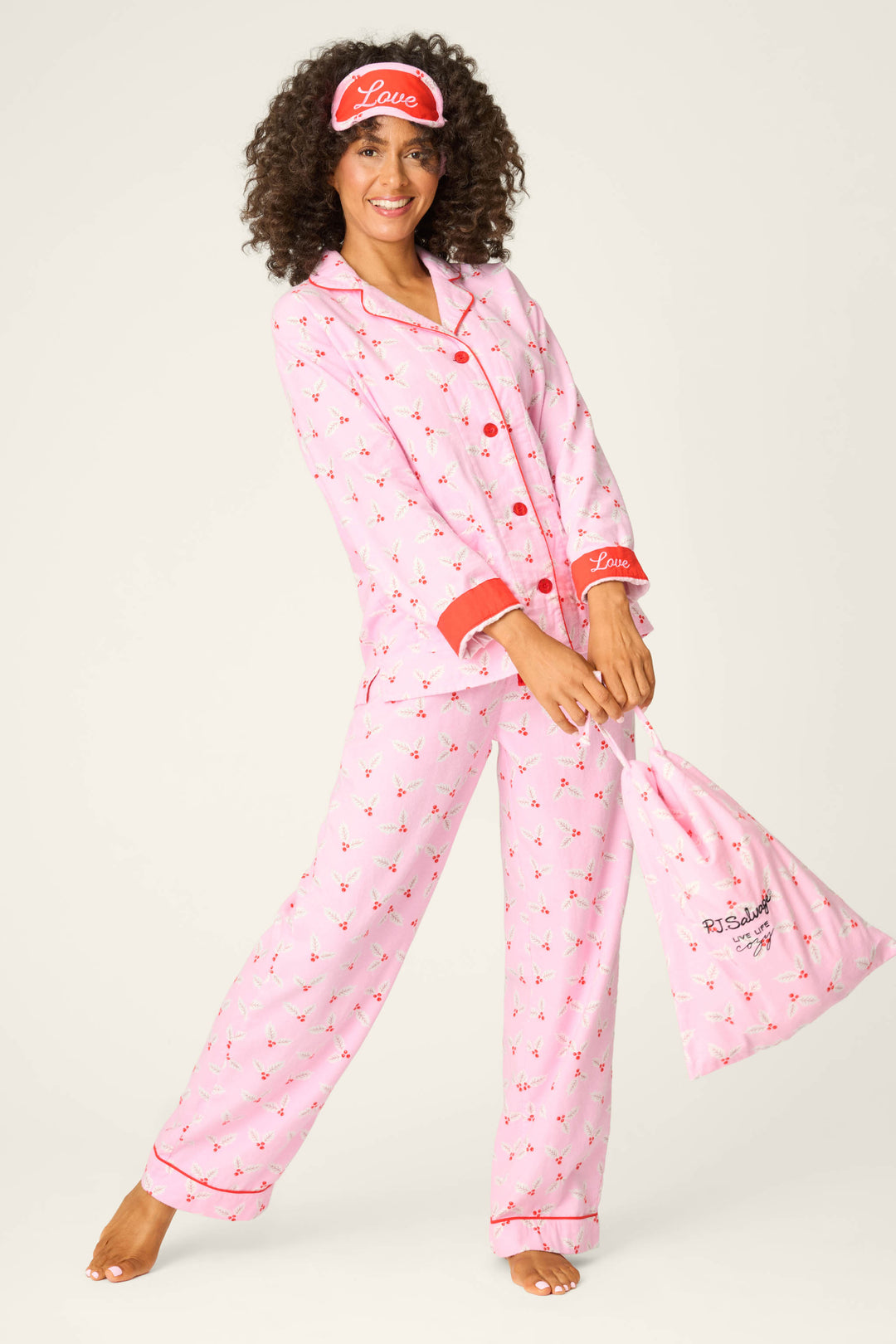 A person stands barefoot in the P.J. Salvage 4-Piece Mistletoe Print Flannel PJ Set, featuring a festive pattern, red accents, matching sleep mask, and a coordinated fabric bag, smiling at the camera against a plain background.