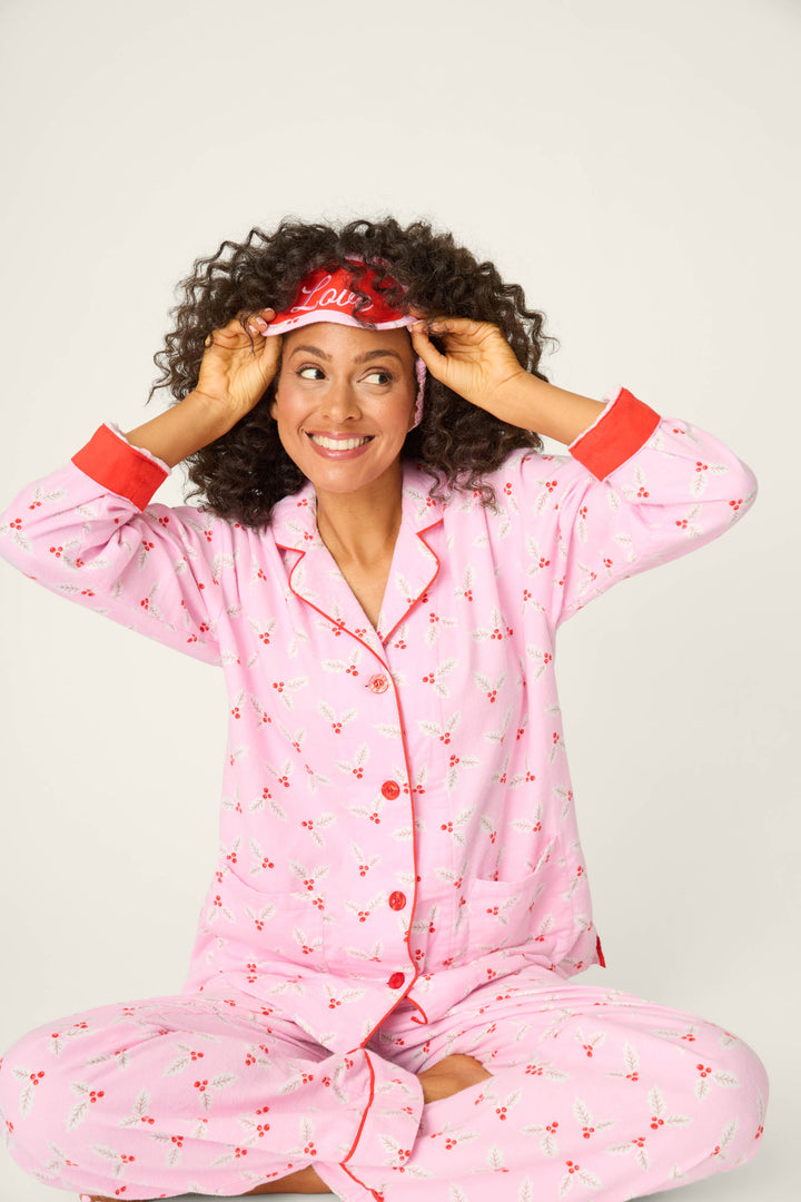 A person in the P.J. Salvage 4-Piece Mistletoe Print Flannel PJ Set sits cross-legged, smiling and adjusting a red “Love” sleep mask, against a plain light background.