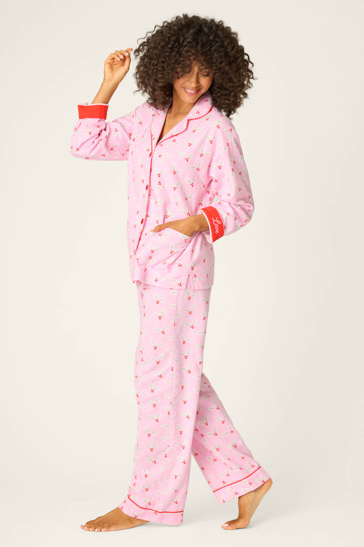 A person with curly hair, barefoot and smiling, wears the P.J. Salvage 4-Piece Mistletoe Print Flannel PJ Set, featuring pink pajama pants and a matching long-sleeve top with red trim and bird pattern, standing on one foot against a light background.