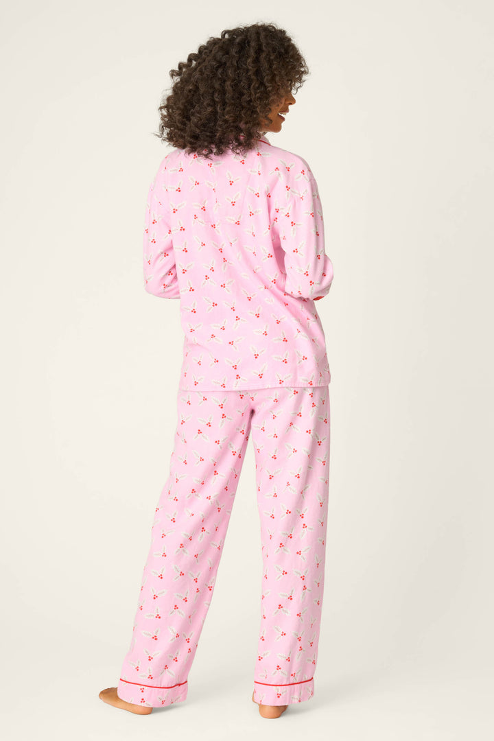 A person with curly hair, barefoot and facing away, wears the P.J. Salvage 4-Piece Mistletoe Print Flannel PJ Set—pink pajamas with a small pattern and red trim—against a plain light background.
