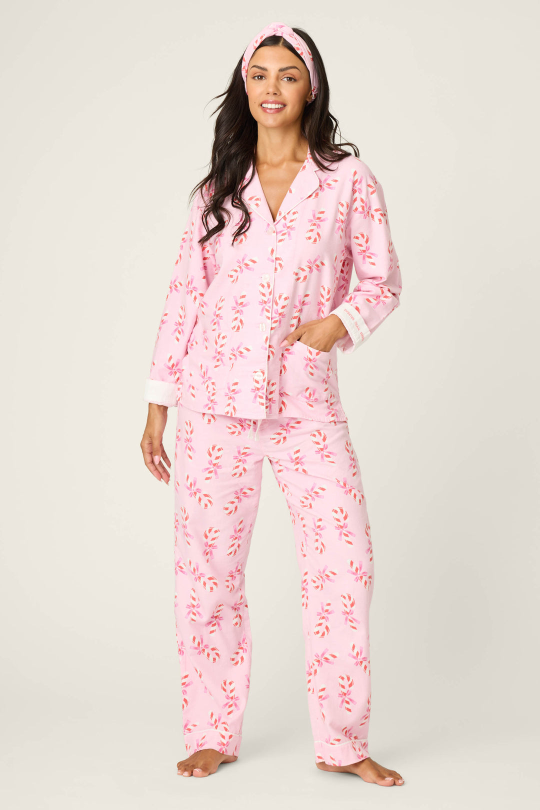 A woman smiles in the P.J. Salvage 3-Piece Candy Cane Print Flannel PJ Set, featuring a matching headband, with one hand in her pocket against a plain light background.