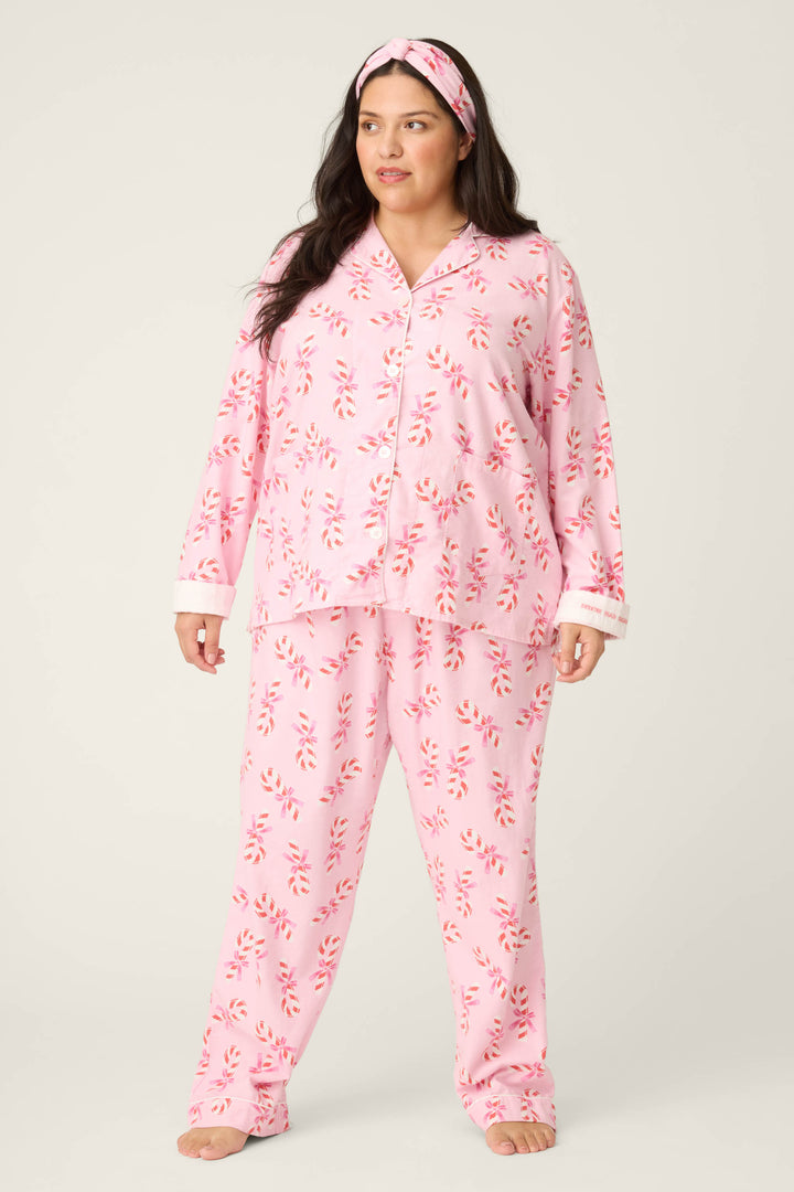 A woman models the P.J. Salvage 3-Piece Candy Cane Print Plus Size Flannel PJ Set, featuring a button-up pajama top, loose-fitting pants, and a matching headband. She stands barefoot against a plain background.
