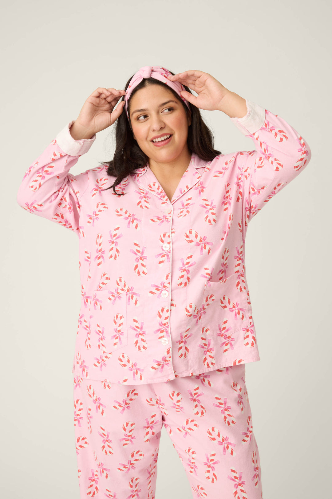 A person smiles at the camera, adjusting their headband while wearing the P.J. Salvage 3-Piece Candy Cane Print Plus Size Flannel PJ Set against a plain background.