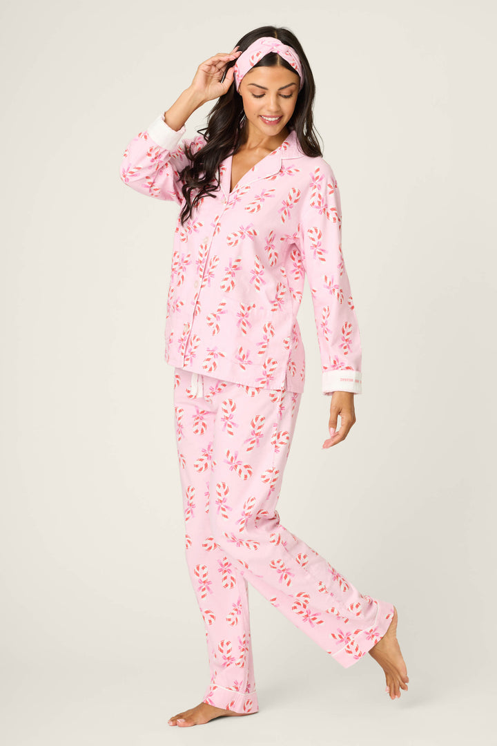 A woman smiles while looking down, standing on one leg in the P.J. Salvage 3-Piece Candy Cane Print Flannel PJ Set with a matching headband, against a plain light background.