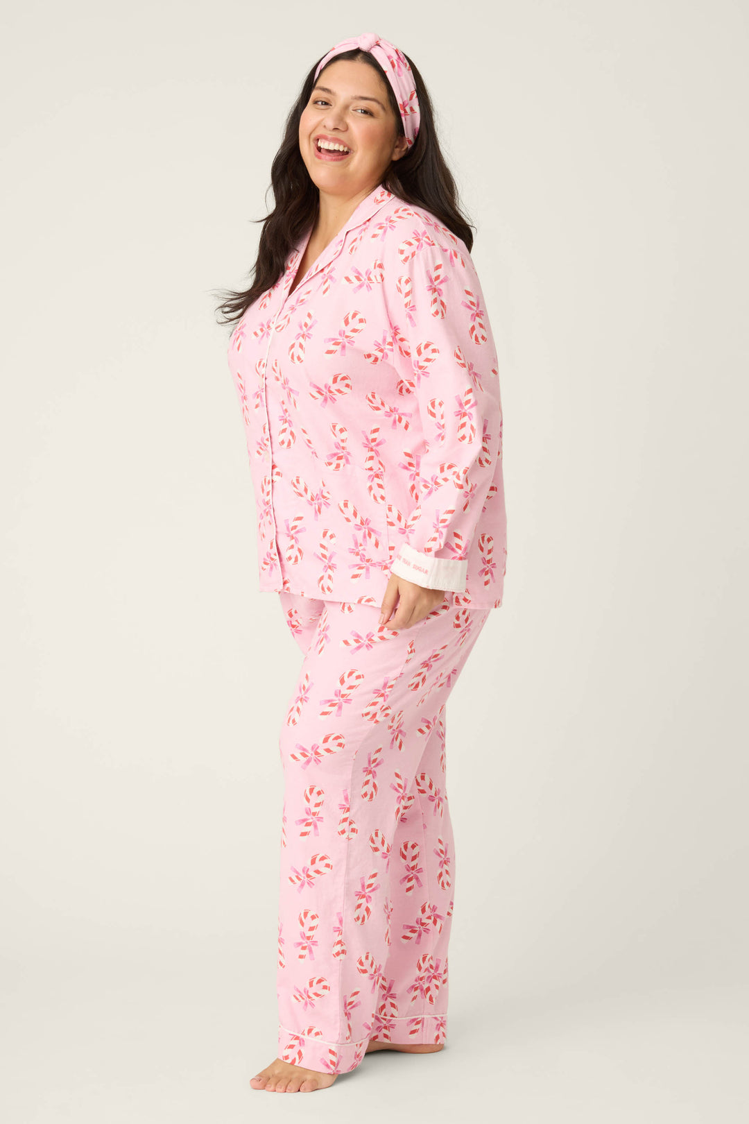 A person smiles barefoot against a plain light background, wearing the P.J. Salvage 3-Piece Candy Cane Print Plus Size Flannel PJ Set and a matching headband.