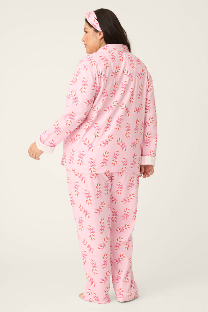 A woman faces away, wearing the P.J. Salvage 3-Piece Candy Cane Print Plus Size Flannel PJ Set, featuring a matching pink long-sleeve top, full-length pants, and a red-and-white patterned headband against a plain light background.