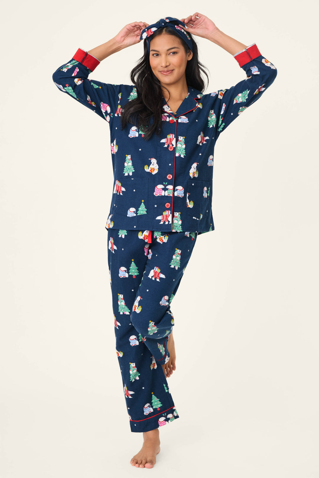 A woman stands barefoot in the P.J. Salvage 3-Piece Bear Print Flannel PJ Set—dark blue pajamas with a festive bear print, red trim, button-up top, long pants, and holds the matching headband above her head.