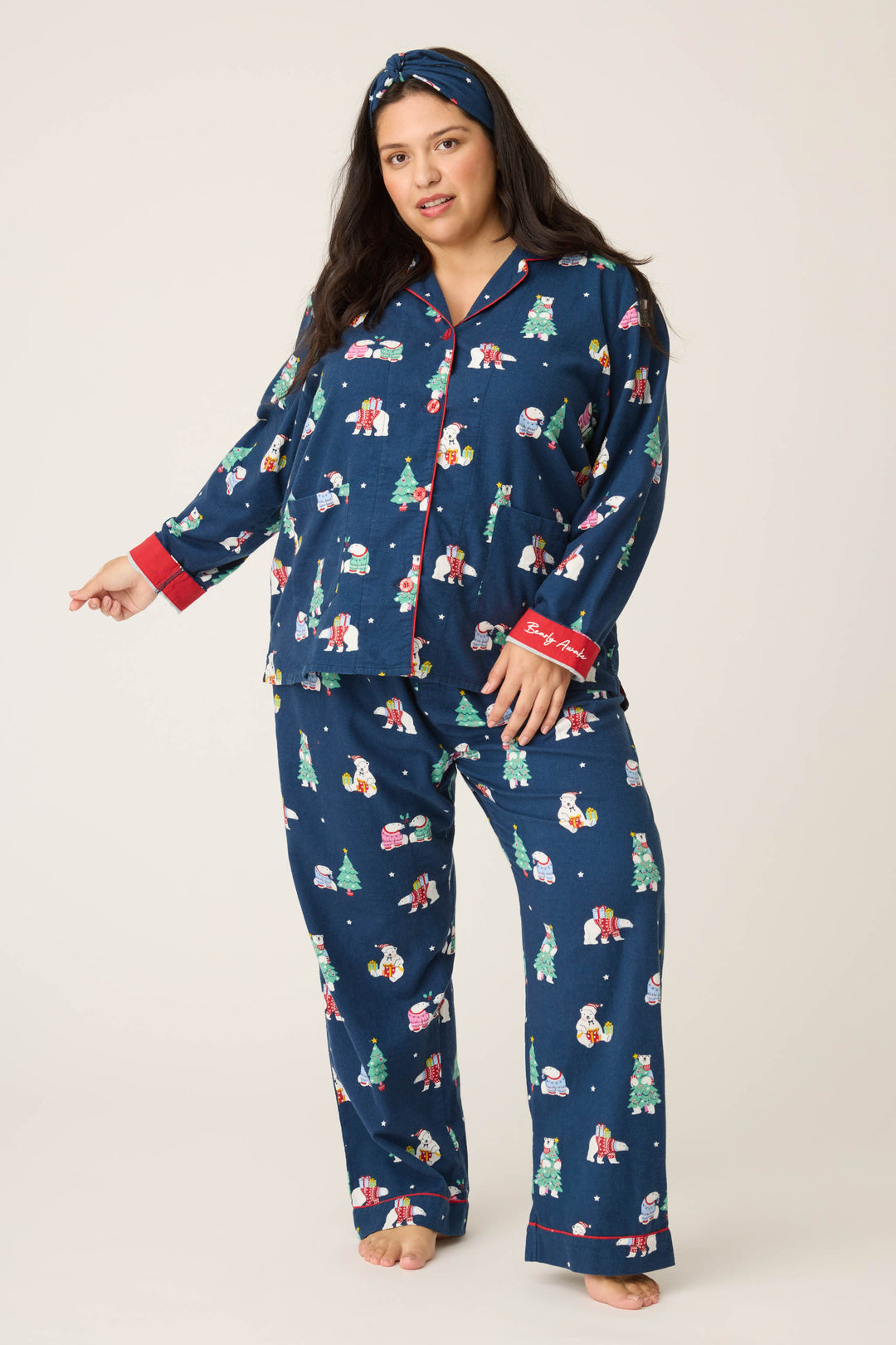 A woman stands barefoot in the P.J. Salvage 3-Piece Bear Print Plus Size Flannel PJ Set, featuring a matching headband and top with red cuffs and buttons. The pajama set has festive prints and a relaxed fit. The background is plain and light-colored.