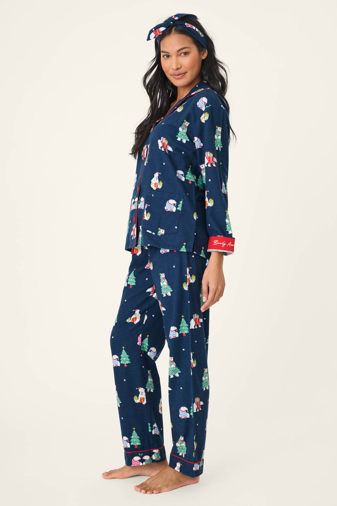 A person stands barefoot in the P.J. Salvage 3-Piece Bear Print Flannel PJ Set, featuring navy blue festive Christmas-themed prints, red cuffs, and a matching headband, against a plain off-white background.