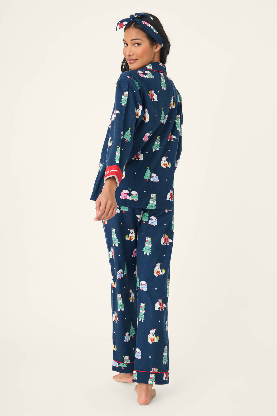 A woman stands barefoot, looking over her shoulder, wearing the P.J. Salvage 3-Piece Bear Print Flannel PJ Set and a colorful, holiday-themed headband with trees and cartoon animals.