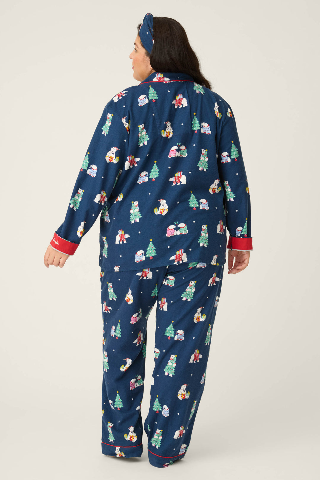 A person with long dark hair, seen from behind, wears the P.J. Salvage 3-Piece Bear Print Plus Size Flannel PJ Set featuring blue holiday-themed patterns with bears, snowmen, and trees, red trim, and a matching blue headband.