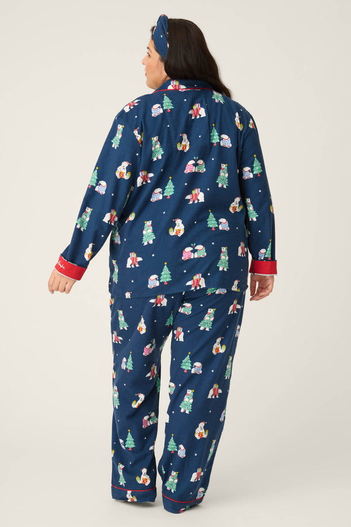 A person with long dark hair, seen from behind, wears the P.J. Salvage 3-Piece Bear Print Plus Size Flannel PJ Set featuring blue holiday-themed patterns with bears, snowmen, and trees, red trim, and a matching blue headband.