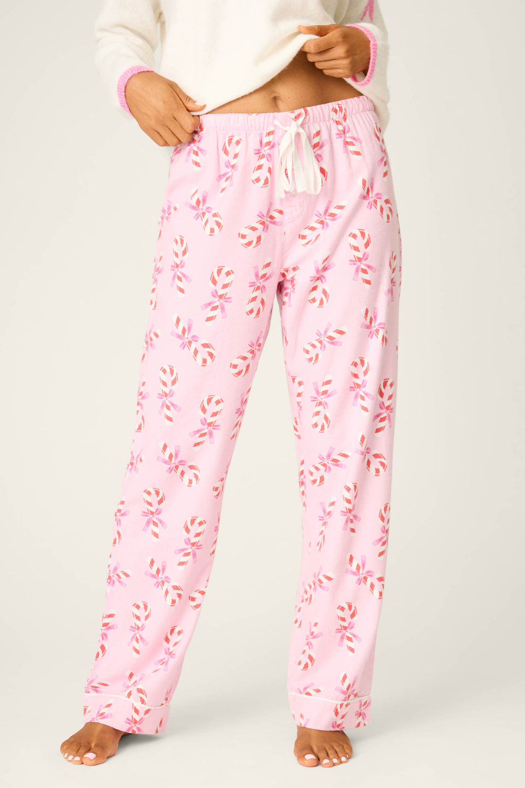 A person, head not visible, stands barefoot wearing P.J. Salvage Candy Cane Print Open Leg Flannel Pants—pink pajama pants featuring a candy cane pattern and a white drawstring.