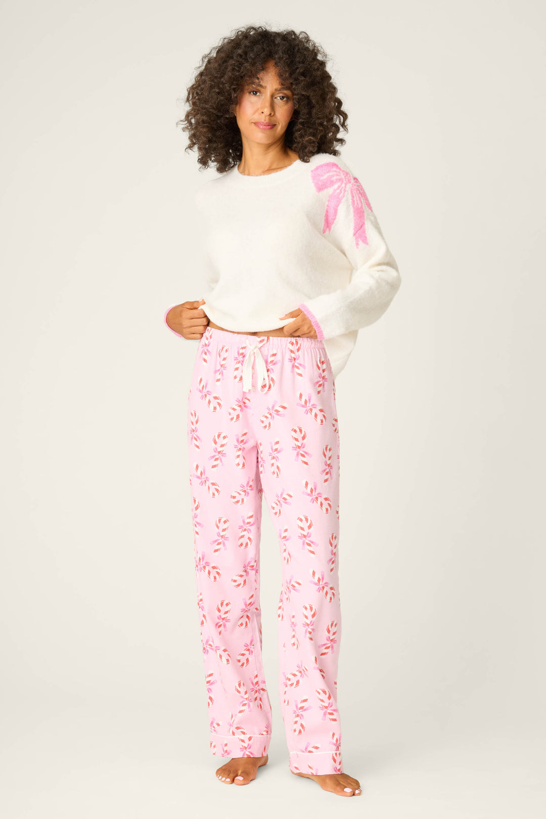 A person with curly hair stands barefoot in a white sweater with a pink bow and P.J. Salvage Candy Cane Print Open Leg Flannel Pants, featuring a red and white candy cane pattern, against a plain light backdrop.