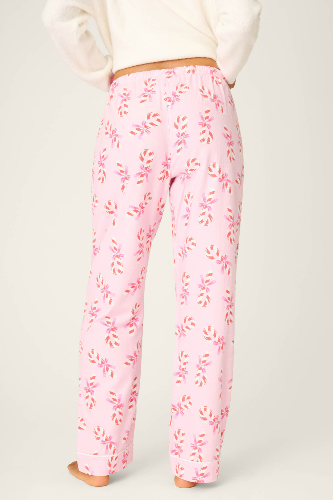 A person stands barefoot on a neutral background, wearing P.J. Salvage Candy Cane Print Open Leg Flannel Pants, shown from the back, featuring a light pink color with a red and white candy cane pattern.