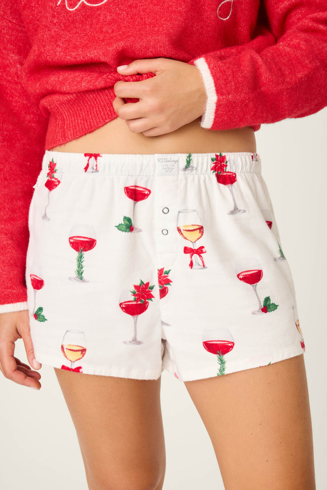 A person wears P.J. Salvage's "Let's Get Toasty Flannel Shorts," featuring festive wine glasses, bows, poinsettias, and holly, paired with a red sweater—ideal for holiday lounging. One hand rests near the shorts’ waistband.