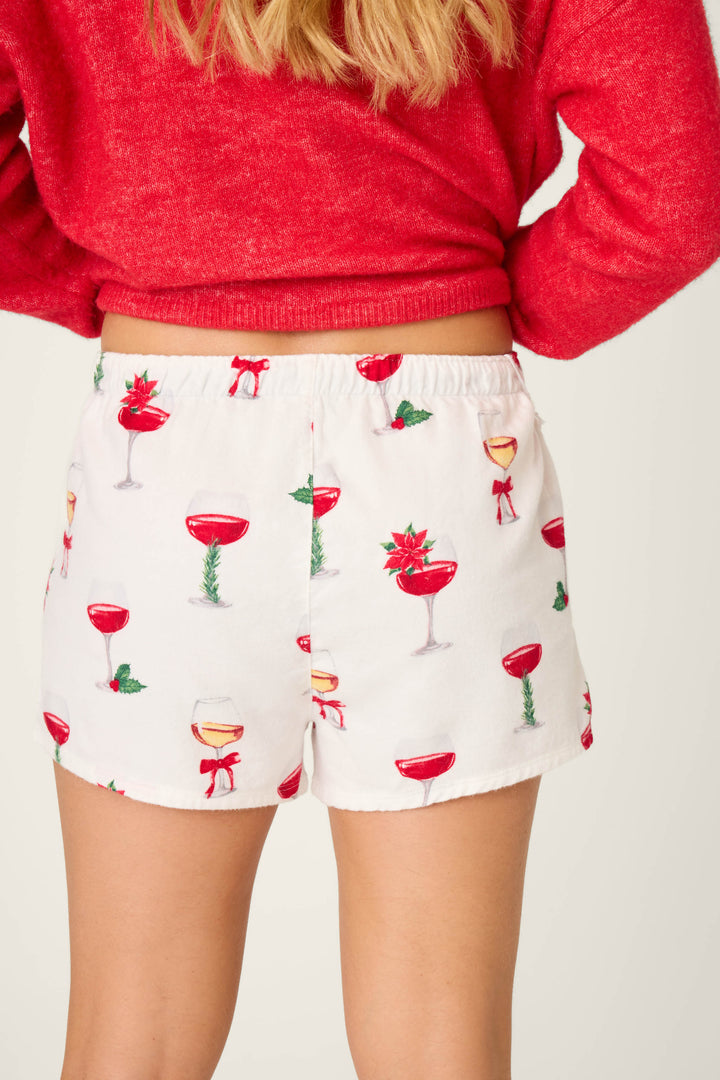 A person wears P.J. Salvage Let's Get Toasty Flannel Shorts, featuring a festive wine glasses pattern, paired with a red sweater. The image captures the waist-down back view, highlighting these perfect holiday lounging shorts.