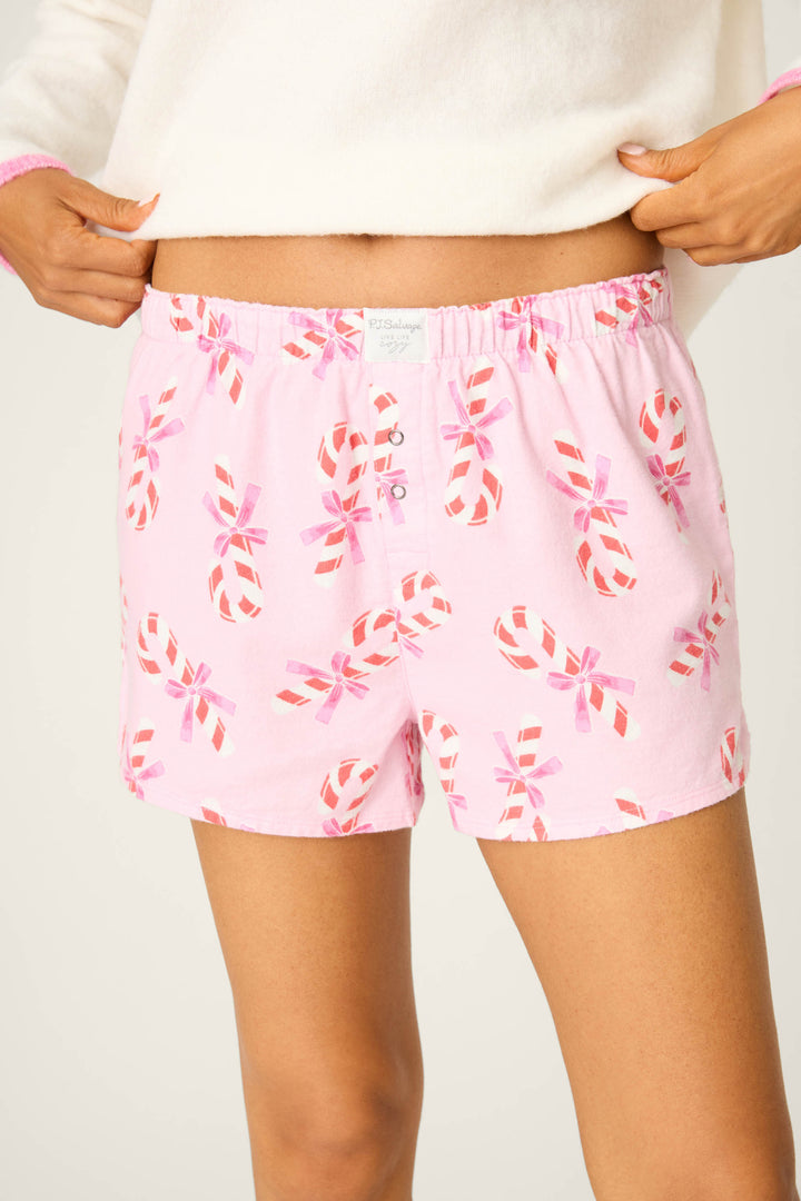 A person wears P.J. Salvage's Sweeter Than Sugar Candy Cane Print Flannel Shorts, shown in pink with red and white candy canes, paired with a fuzzy white top. The cropped photo highlights the cozy holiday shorts on the torso and upper legs.