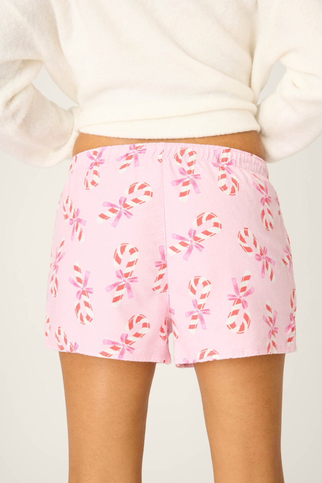 A person is shown from the back wearing P.J. Salvage Sweeter Than Sugar Candy Cane Print Flannel Shorts with pink bows and a white long-sleeve top, perfect for a cozy holiday look.