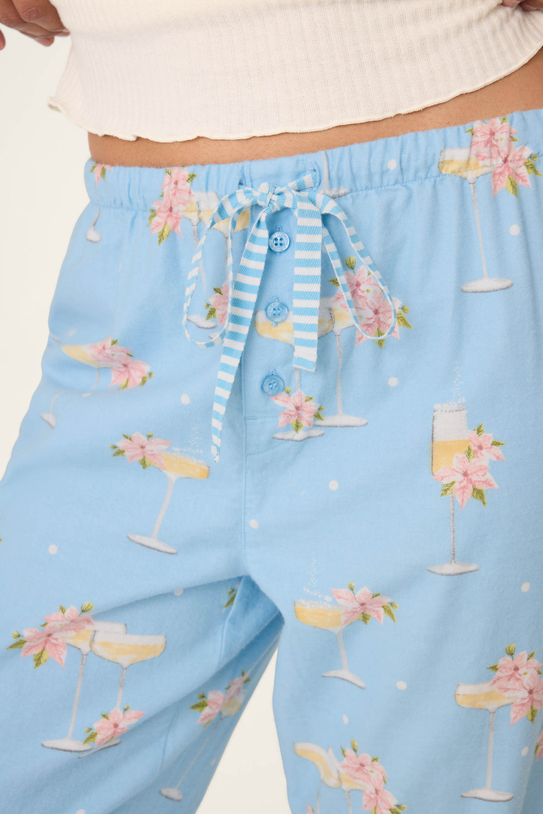 A close-up of the P.J. Salvage Fizz the Season Open Leg Flannel Pants features light blue fabric with champagne glass and pink flower print, a white and blue striped drawstring, blue buttons, and part of a white ribbed top waistband.
