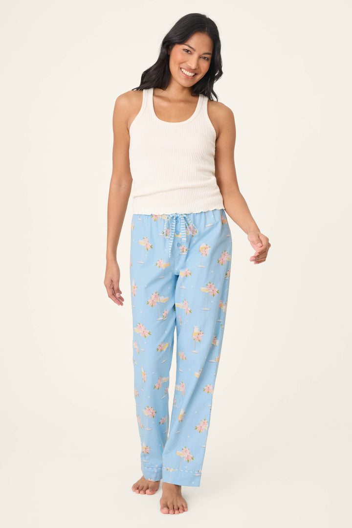 A person with long dark hair smiles barefoot against a plain background, wearing a white ribbed tank and P.J. Salvage Fizz the Season Open Leg Flannel Pants in light blue with peach and floral print.