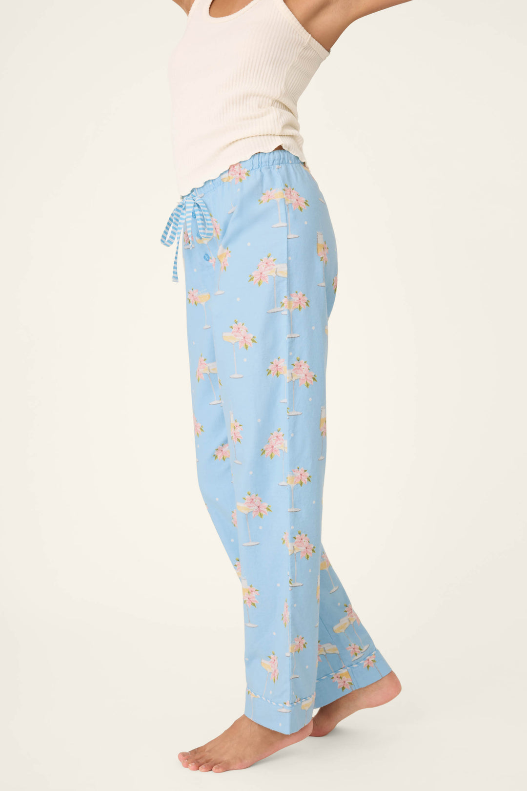 A person wears P.J. Salvage's Fizz the Season Open Leg Flannel Pants—light blue with a pink palm tree print—and a white ribbed tank top, standing barefoot against a plain background. The image is cropped above their nose.