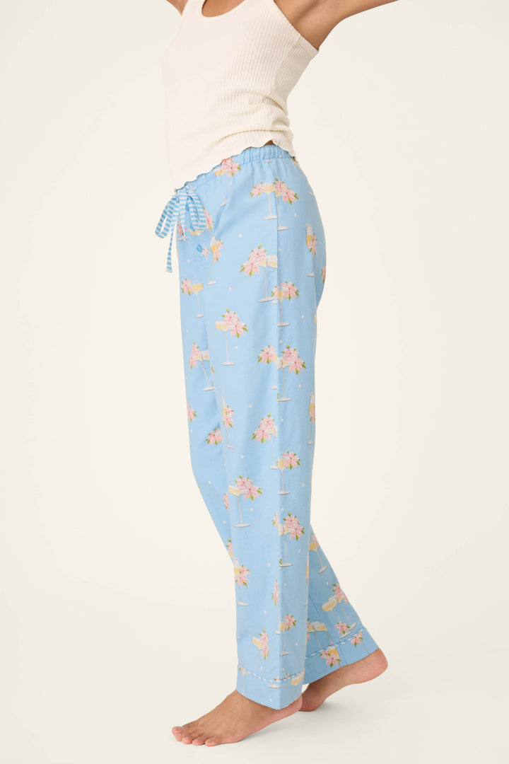 A person wears P.J. Salvage's Fizz the Season Open Leg Flannel Pants—light blue with a pink palm tree print—and a white ribbed tank top, standing barefoot against a plain background. The image is cropped above their nose.