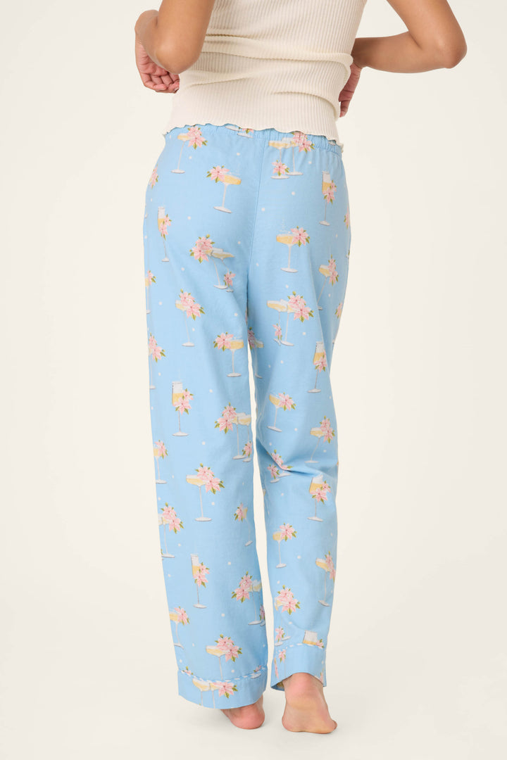 A person stands barefoot, facing away, wearing P.J. Salvage's Fizz the Season Open Leg Flannel Pants featuring a pink and yellow floral cocktail print with a white ribbed tank top against a plain, light background.