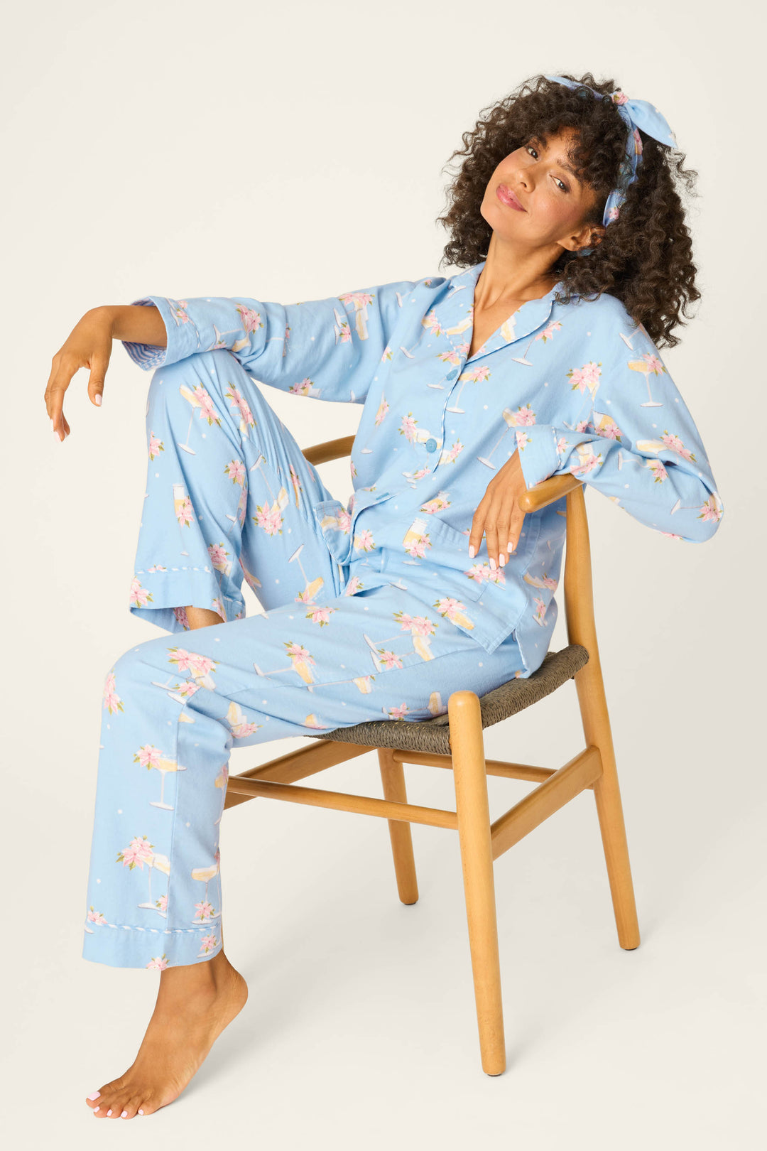 A person with curly hair and a matching hairband sits on a wooden chair, wearing the P.J. Salvage Fizz The Season 3-Piece Flannel PJ Set in light blue floral. One leg is raised on the chair while the other rests on the floor.