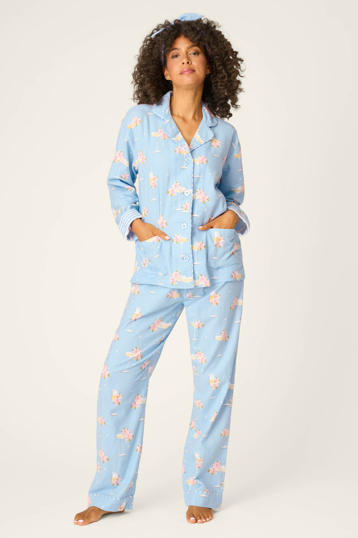 A woman with curly hair stands barefoot in the P.J. Salvage Fizz The Season 3-Piece Flannel PJ Set—light blue, long-sleeved, button-up pajamas with a pink floral print, hands in pockets, against a plain, light background.