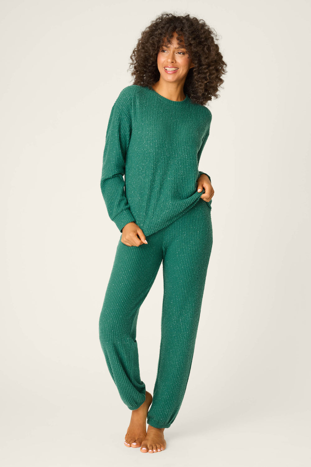 A person with curly hair smiles barefoot against a plain background, holding the hem of a green P.J. Salvage SOLID PJ SET FIRESIDE SPARKLE SETS top and matching pants.