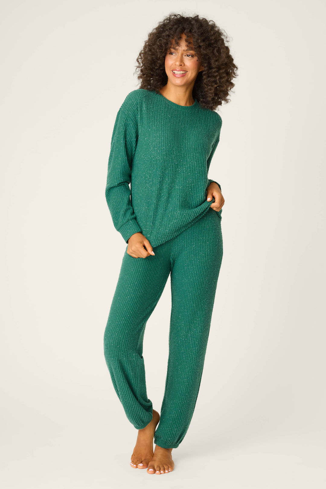 A person with curly hair smiles barefoot against a plain background, holding the hem of a green P.J. Salvage SOLID PJ SET FIRESIDE SPARKLE SETS top and matching pants.