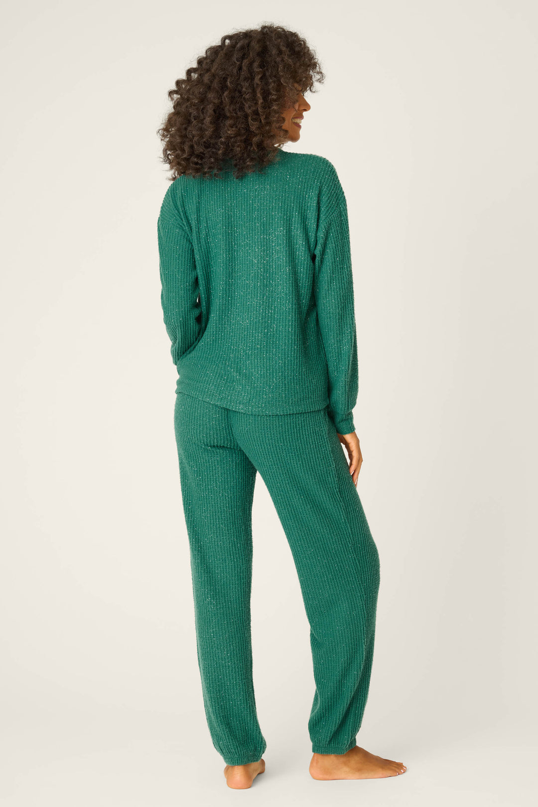 A person with curly hair stands barefoot on a plain background, facing away from the camera, wearing the P.J. Salvage SOLID PJ SET FIRESIDE SPARKLE SETS—a matching green textured long-sleeve top and pants set.
