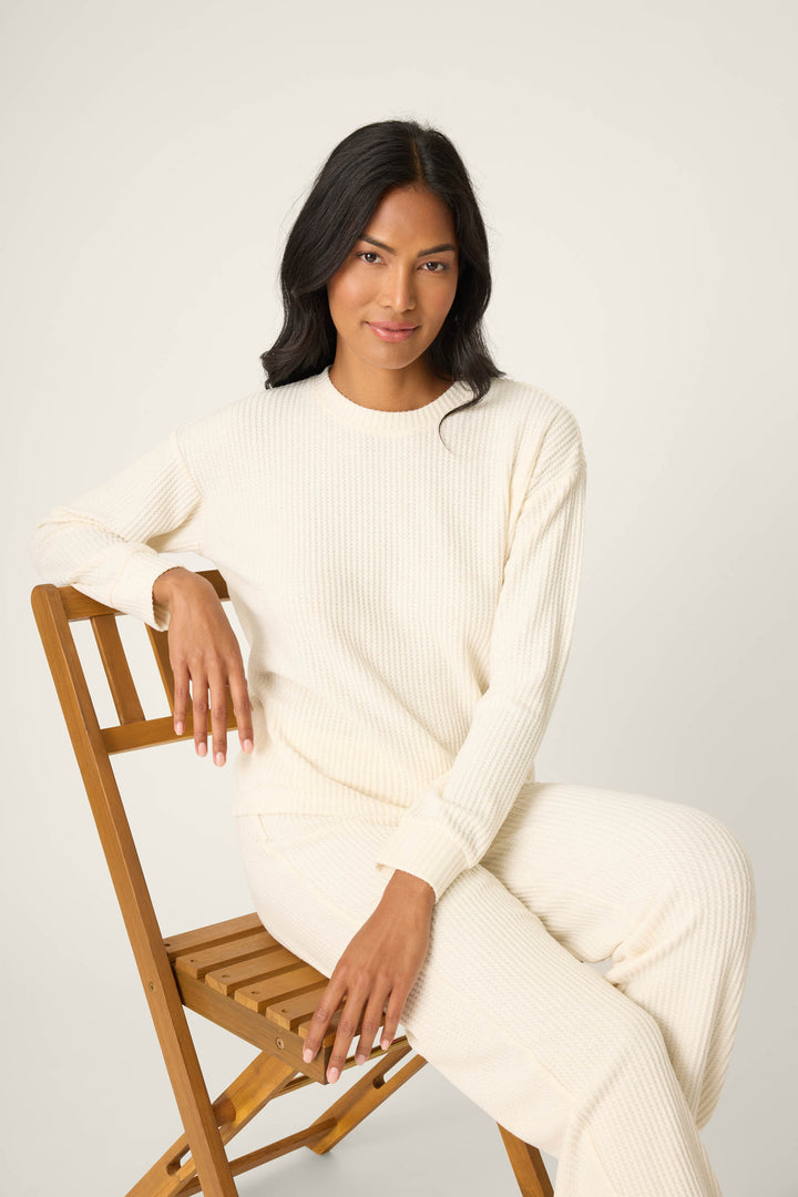 A person with long, dark hair sits on a wooden folding chair, wearing the cream-colored SOLID PJ SET FIRESIDE SPARKLE SETS by P.J. Salvage, against a plain light background.