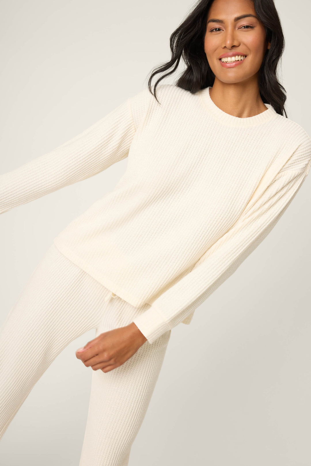 A person wearing the P.J. Salvage SOLID PJ SET FIRESIDE SPARKLE SETS, featuring a cream-colored, long-sleeve, waffle-knit top and matching pants, stands smiling against a plain light background.