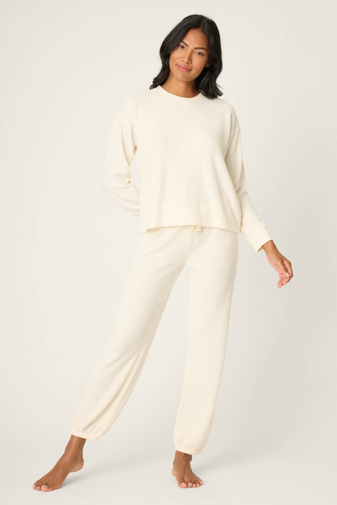 A woman stands barefoot against a plain background, wearing the P.J. Salvage SOLID PJ SET FIRESIDE SPARKLE SETS—matching cream-colored, long-sleeve ribbed top and pants loungewear—with one hand behind her back and a neutral expression.