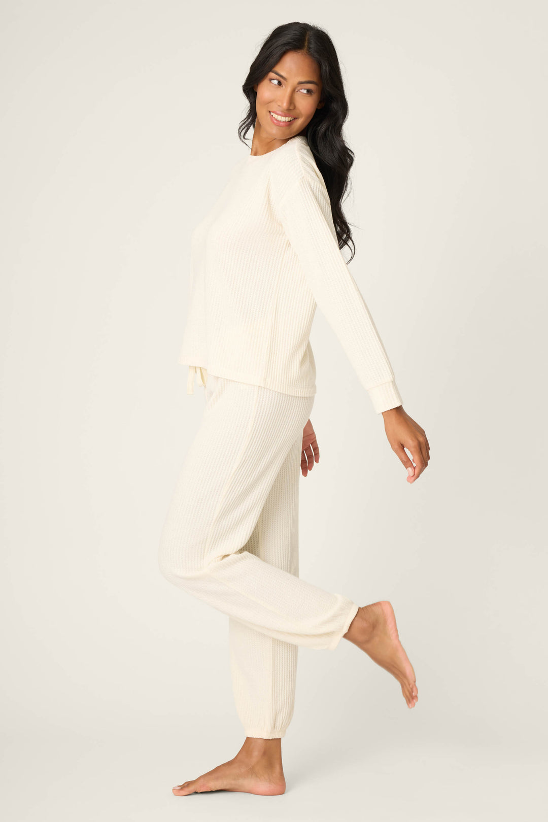 A woman with long dark hair wears the SOLID PJ SET FIRESIDE SPARKLE SETS by P.J. Salvage. She smiles, looking over her shoulder, standing on one leg against a light, plain background.