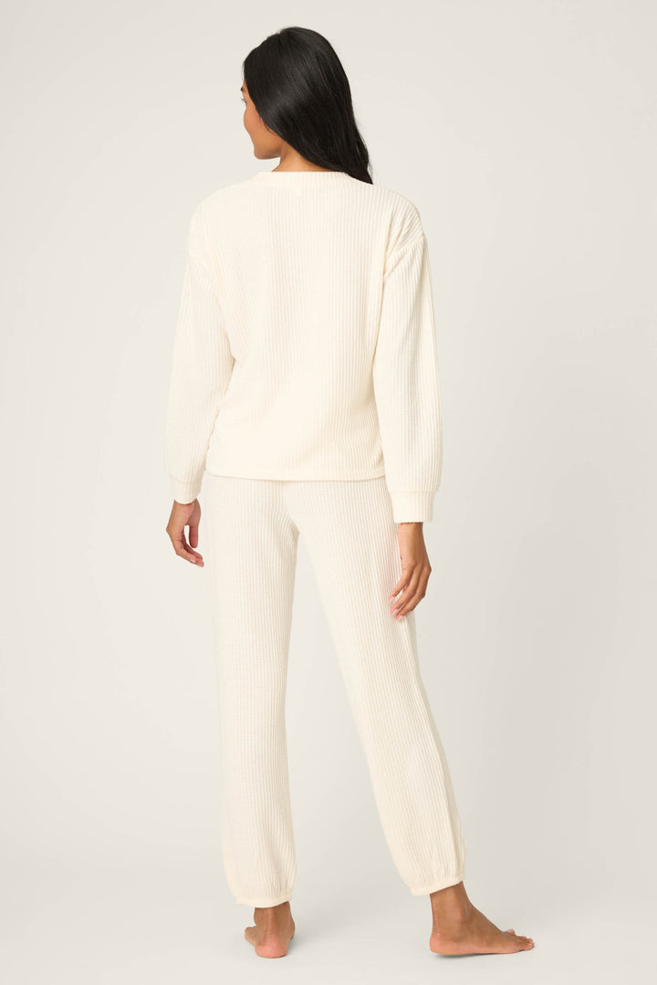 A person with long dark hair stands barefoot facing away, wearing the cream-colored SOLID PJ SET FIRESIDE SPARKLE SETS by P.J. Salvage—featuring a long-sleeve ribbed top and matching ribbed pants—against a plain, light background.