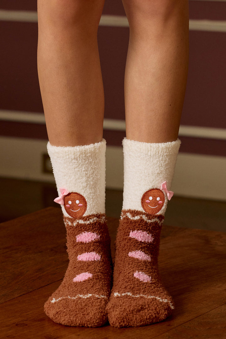 A person wears P.J. Salvage GINGERBREAD MAN SOCKS SKI JAMMIE CLASSICS—fuzzy brown and white socks with pink-striped gingerbread faces and bows—while standing on a wooden floor against a maroon background with white trim.