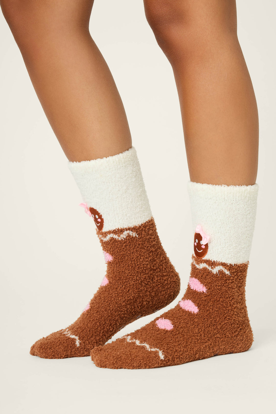 A person is shown from the lower legs down wearing P.J. Salvage GINGERBREAD MAN SOCKS SKI JAMMIE CLASSICS—fuzzy brown and white socks featuring a smiling gingerbread cookie design with pink icing and a pink bow.