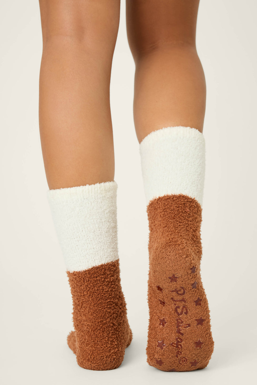 A person stands on a light surface wearing P.J. Salvage GINGERBREAD MAN SOCKS SKI JAMMIE CLASSICS, which have brown textured, non-slip soles with printed text and stars and white fuzzy uppers.