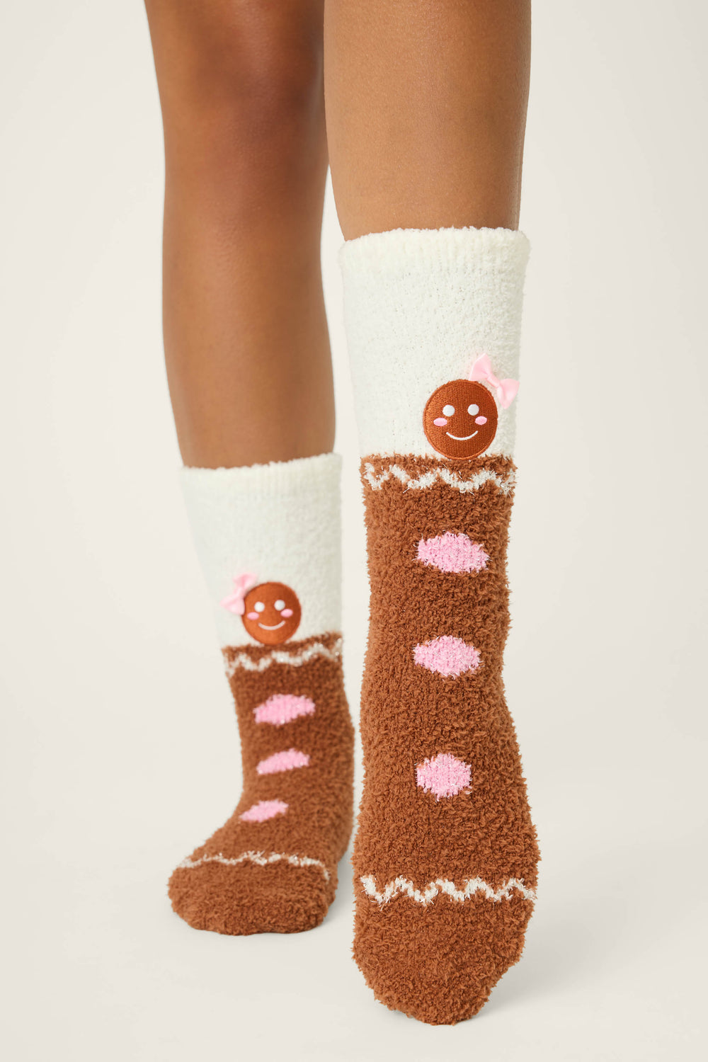 Someone wears P.J. Salvage GINGERBREAD MAN SOCKS SKI JAMMIE CLASSICS—fuzzy white and brown socks with pink dots, a gingerbread cookie face, and a pink bow—against a plain, light background.